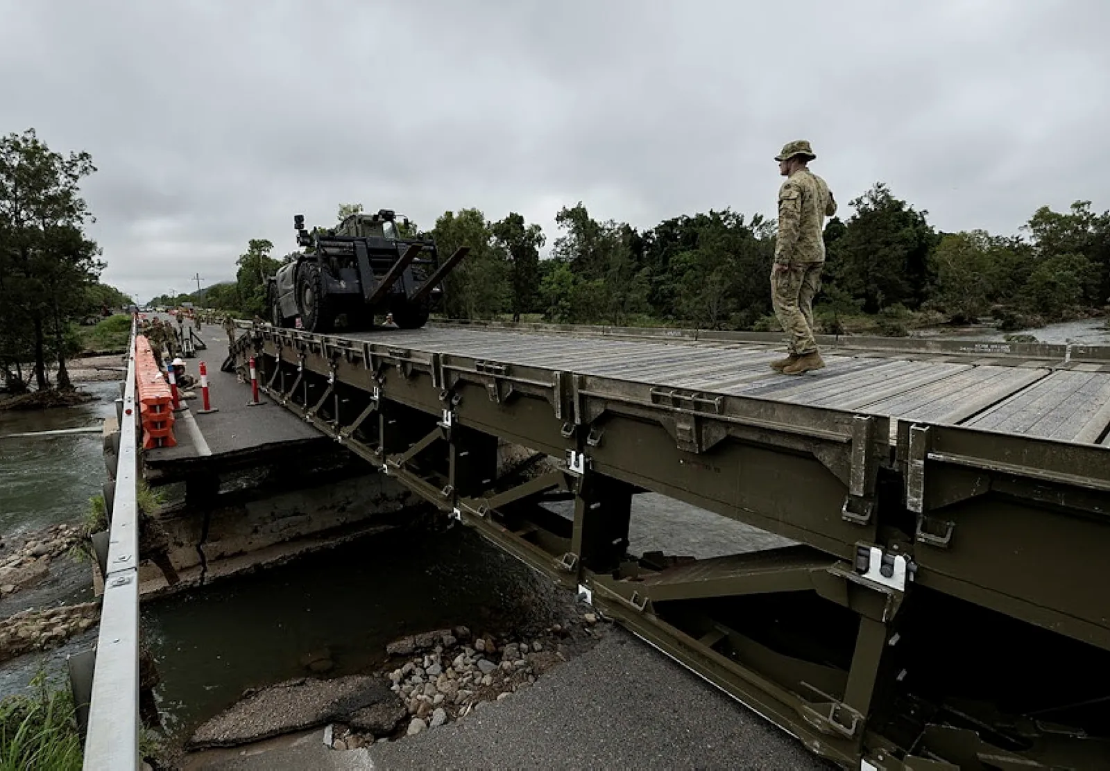 A soldier walks a deployed military bridge as an armoured vehicle approaches the crossing